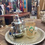 Silver teapot and colourful glass on a silver tray. Colourful market stalls are in the background