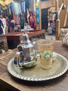 Silver teapot and colourful glass on a silver tray. Colourful market stalls are in the background