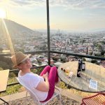 Woman in pink trousers, white top, and a hat is looking at the camera over her shoulder. In front of her is a hillside city view and on the table next to her is a glass of beer.