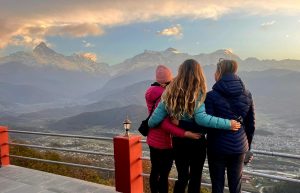 Three women, facing away from the camera and towards the Annapurna range, stand with arms interlinked behind their backs